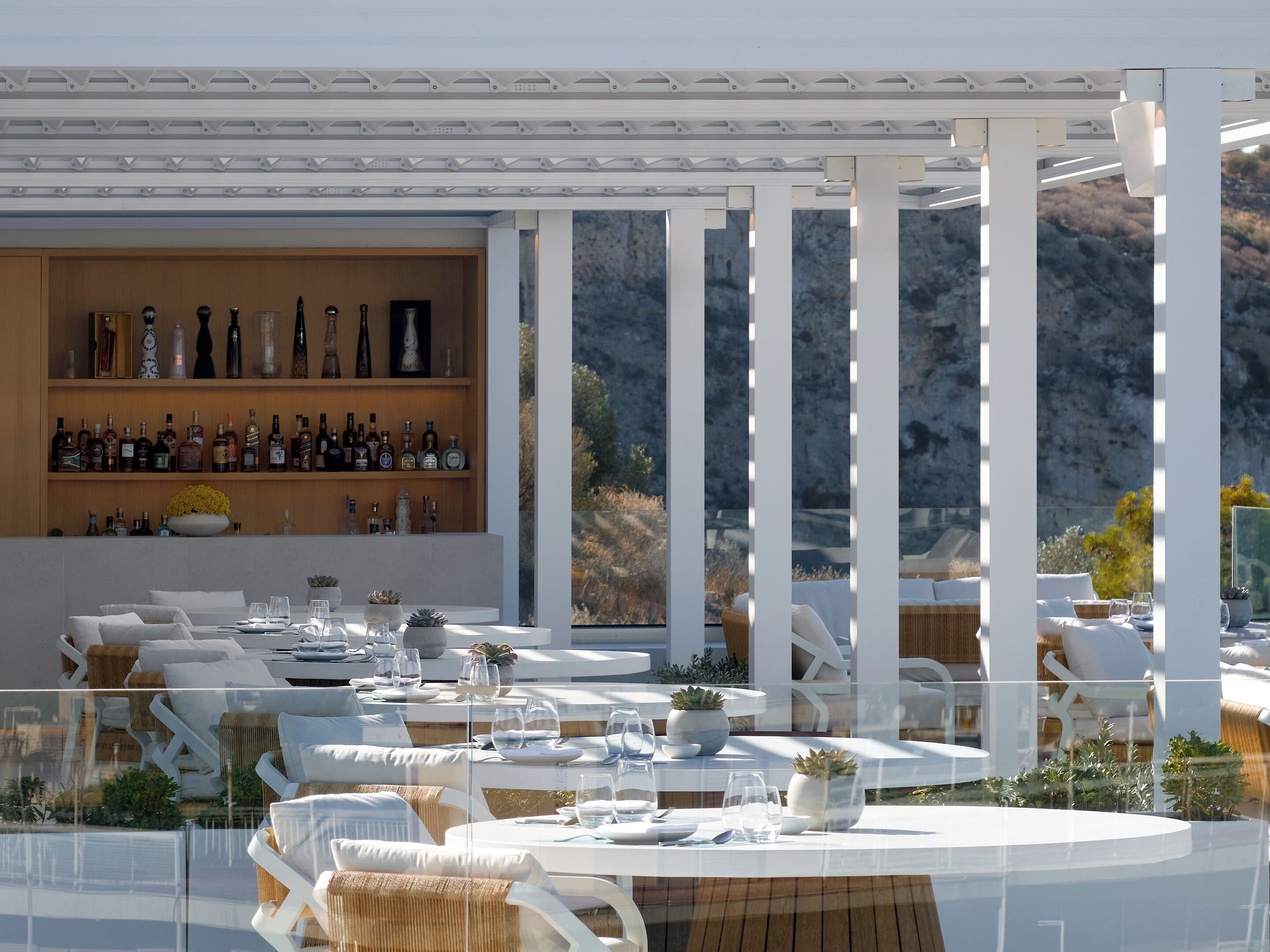 Un área de comedor al aire libre moderna, con mobiliario blanco, una barra con estantes llenos de botellas y vistas del paisaje circundante.
