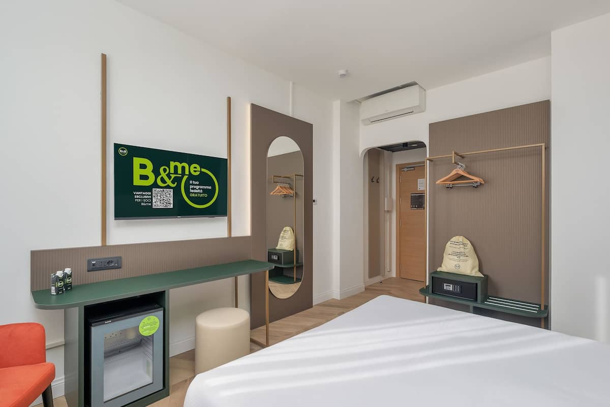 A hotel room with a bed, a desk with a computer, a mini-fridge, a mirror, a wardrobe, and a hanging clothes rack.