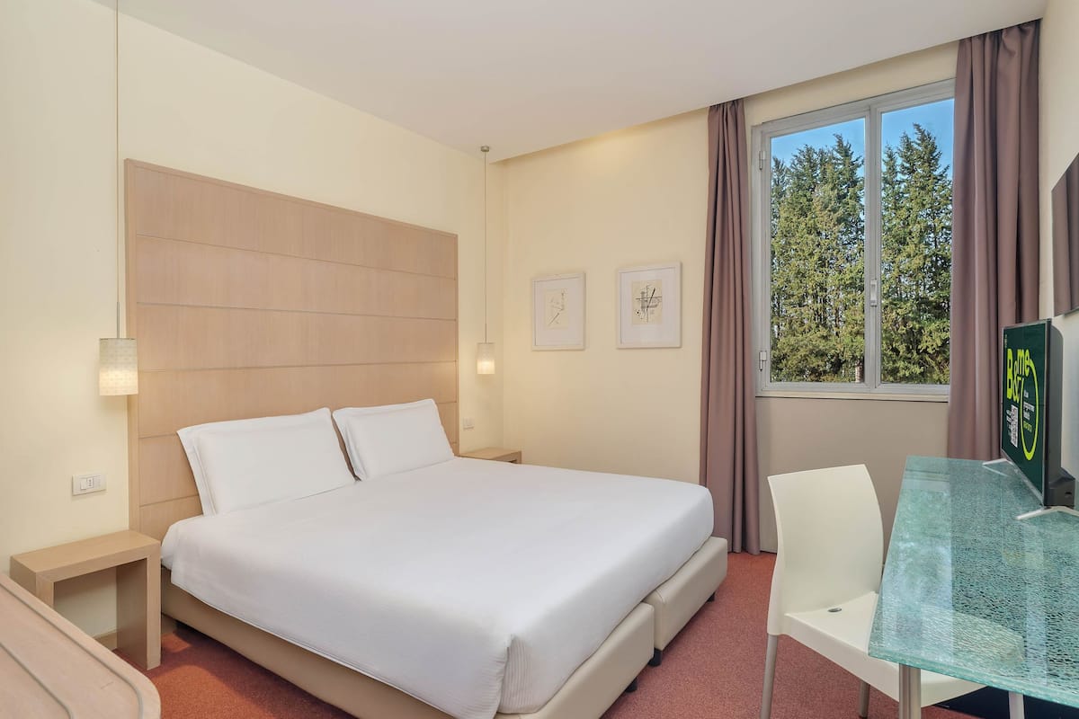 A hotel room with a bed, bedside tables, a desk with a chair, a window with curtains, and framed pictures on the wall.