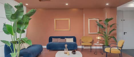Un salon moderne avec un canapé bleu, une table basse et deux fauteuils. De grandes plantes en pot et des tableaux encadrés ornent les murs.