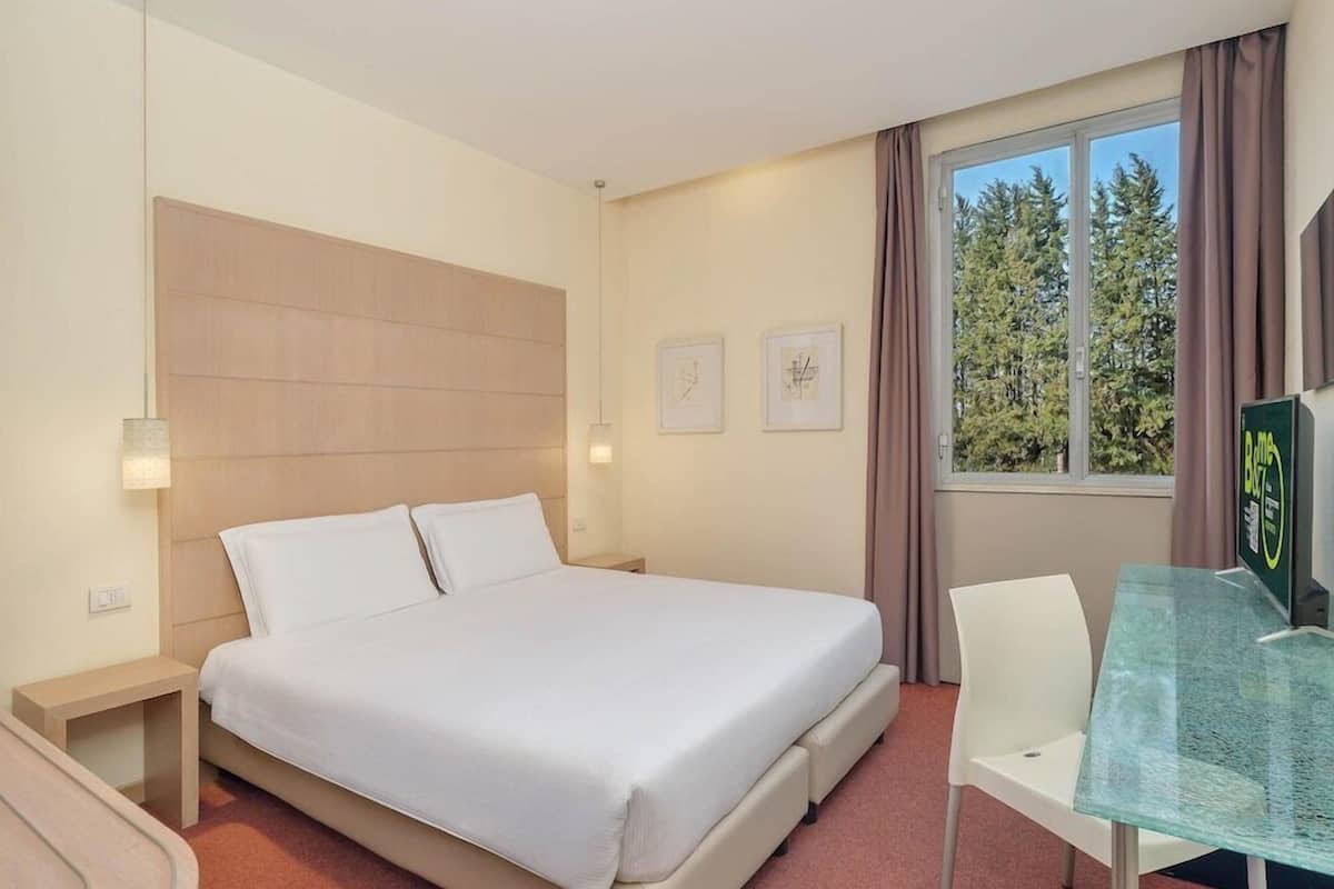 A hotel room with a bed, bedside tables, a desk with a chair, a window with a view of trees, and wall art.