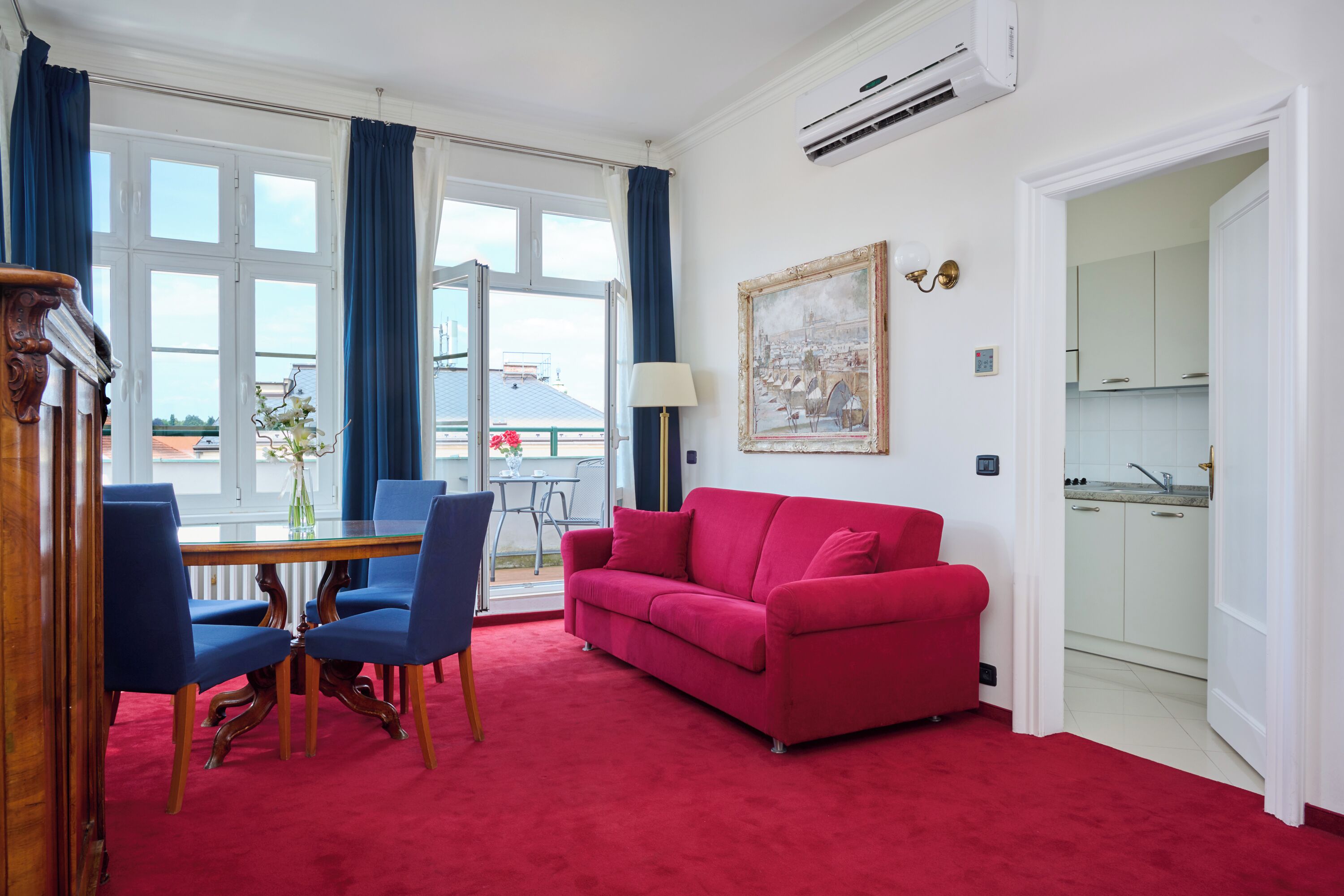 A room with a red carpet, a red sofa, a dining table with blue chairs, and a view of the outside through large windows with blue curtains.