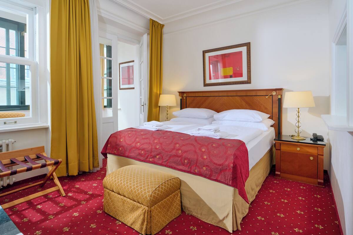 Una habitación de hotel con una cama grande, dos mesitas de noche, un banco de madera, un pequeño reposapiés y una ventana con cortinas amarillas.