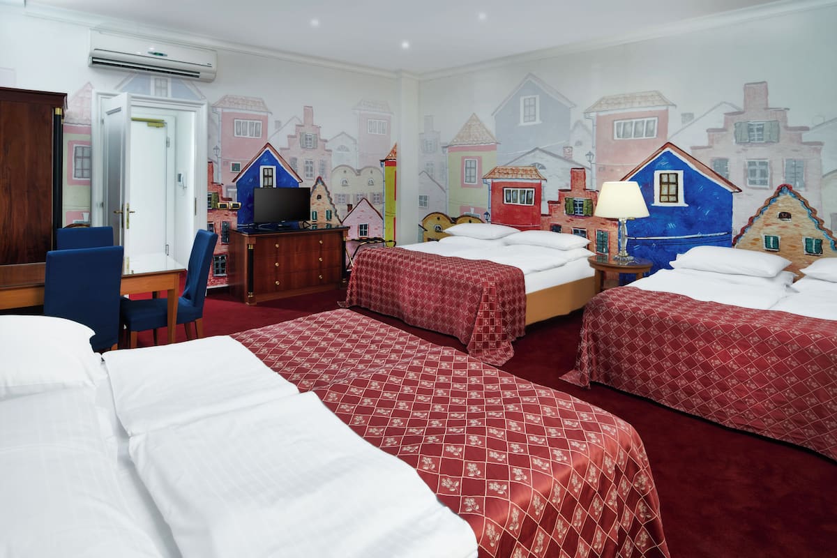Habitación de hotel con dos camas, un escritorio con silla, un televisor y un mural en la pared con casas.