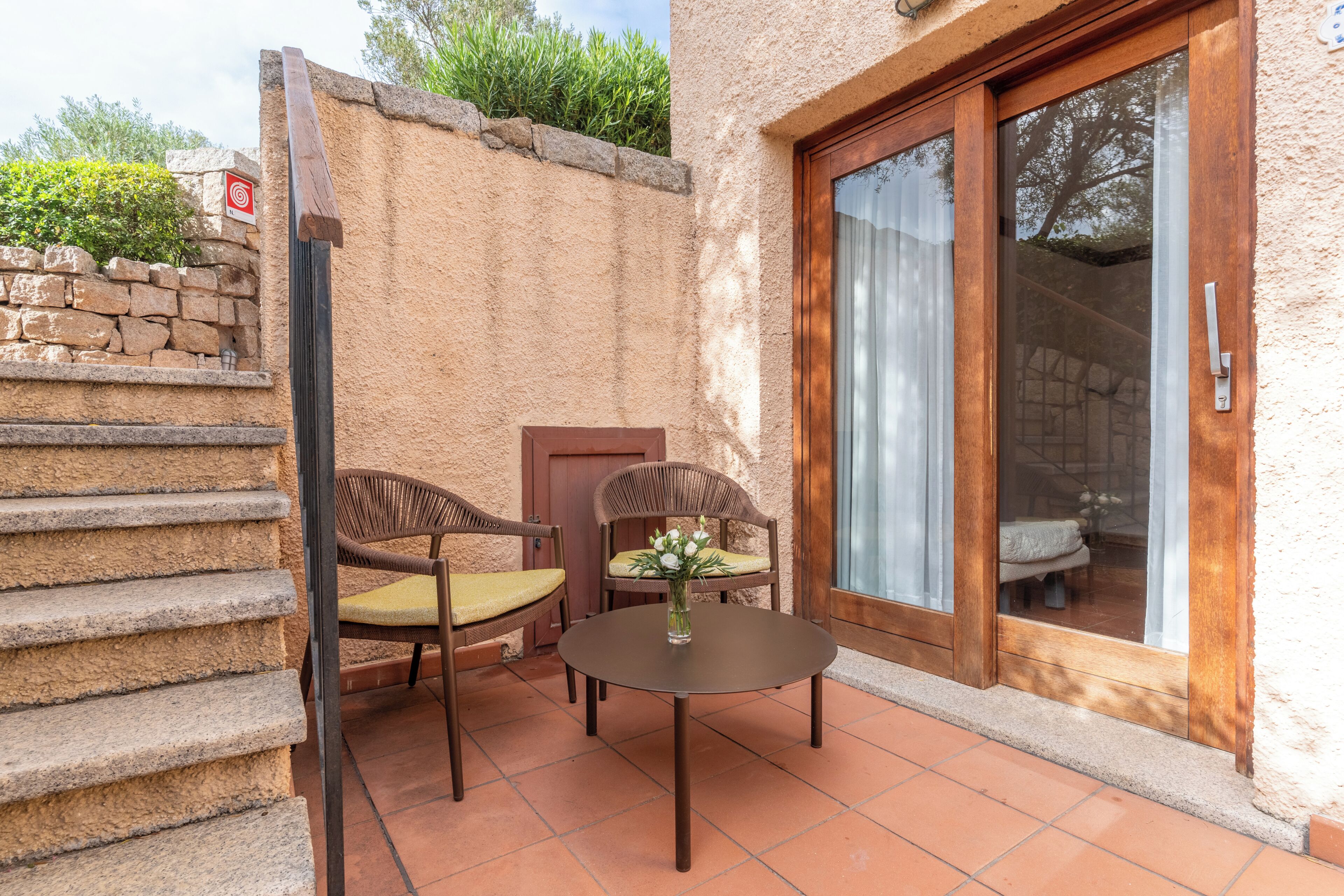 A patio with a round table and two wicker chairs, a small vase with flowers, and a sliding glass door.