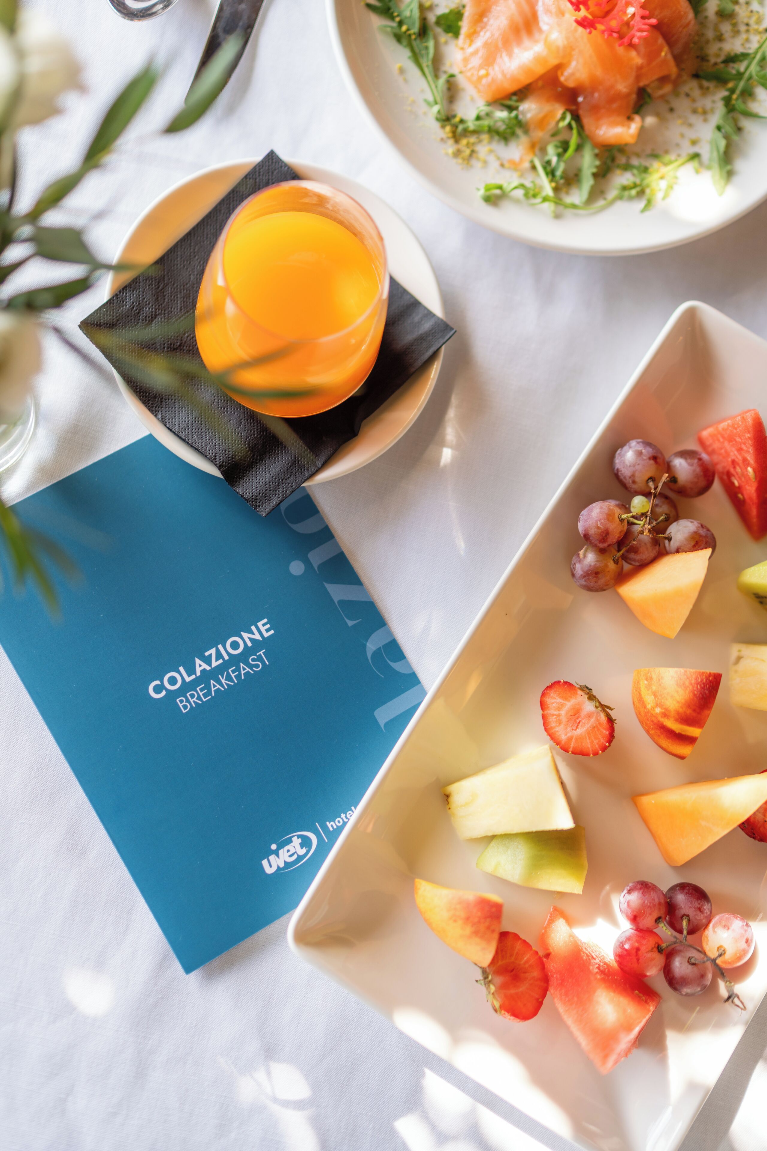 Une table mise pour le petit-déjeuner avec une assiette de fruits, un verre de jus d’orange et un menu intitulé « Colazione Breakfast ».
