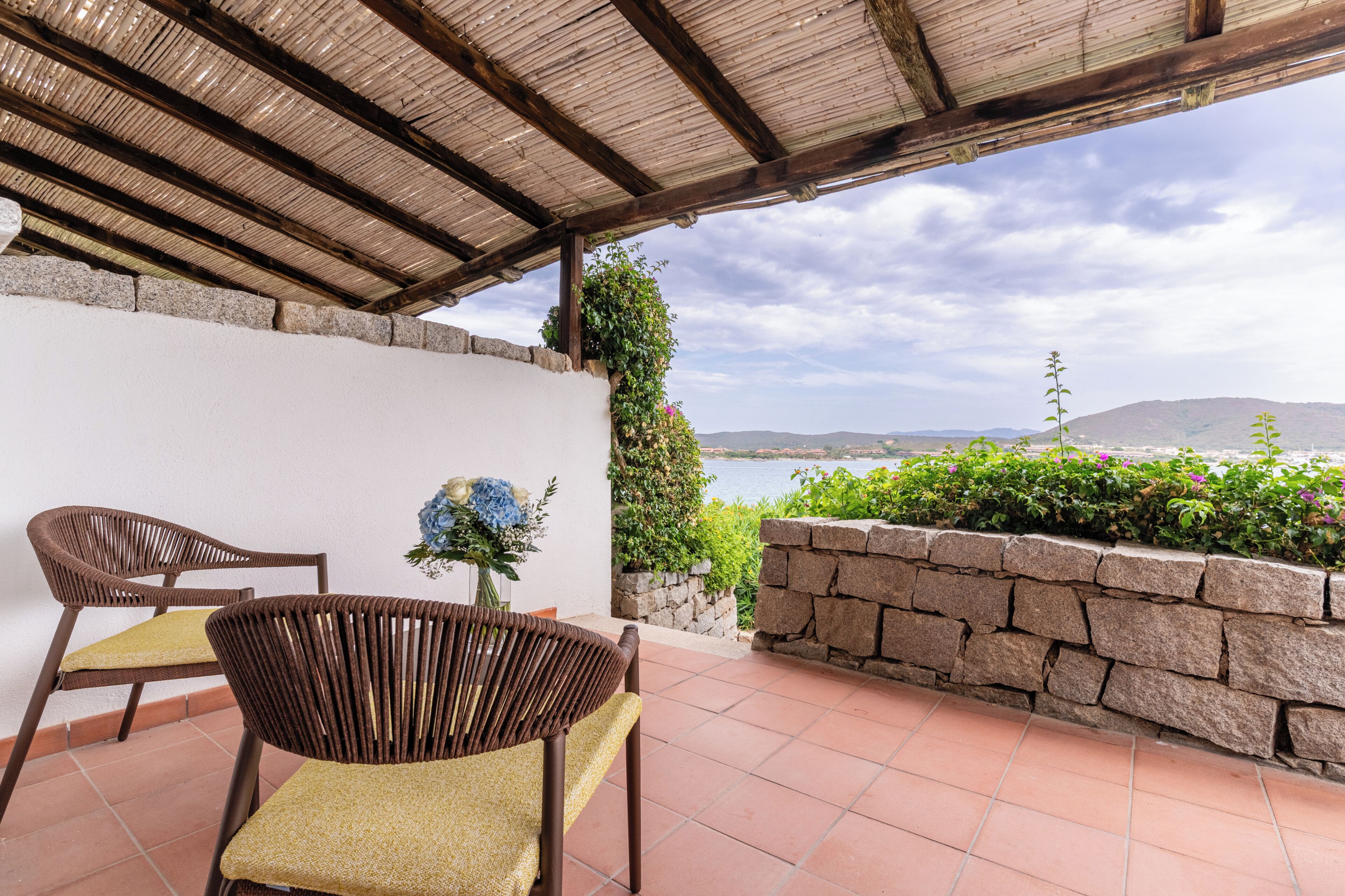 Um alpendre coberto com duas cadeiras de vime, uma mesa e vista para o mar e as colinas.