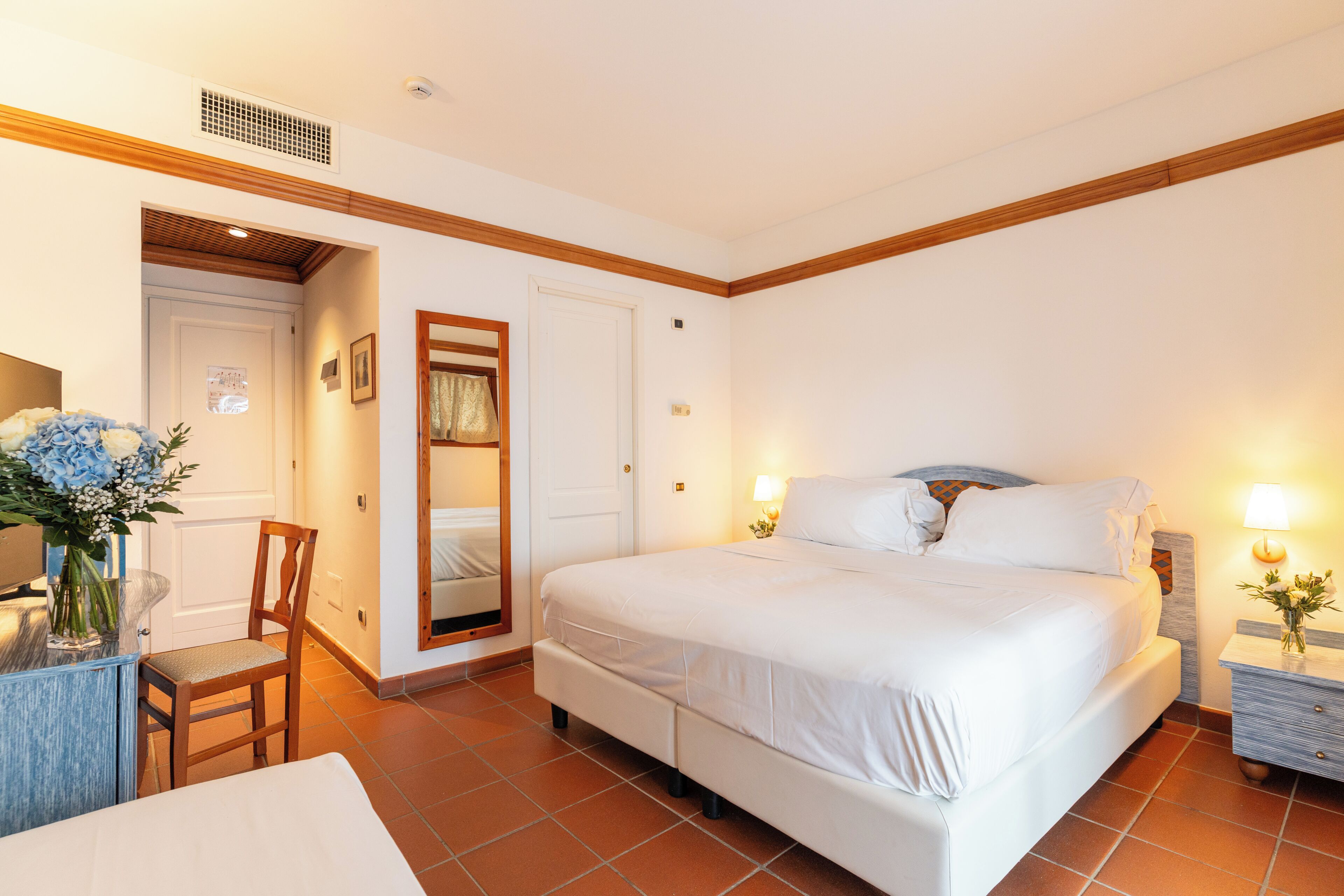 A hotel room with a large bed, a desk with a chair, a mirror, and a nightstand with a lamp.