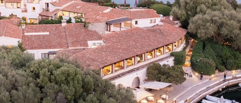 A coastal building with a terrace overlooking the sea, a dock with boats, and a pathway leading to the entrance.