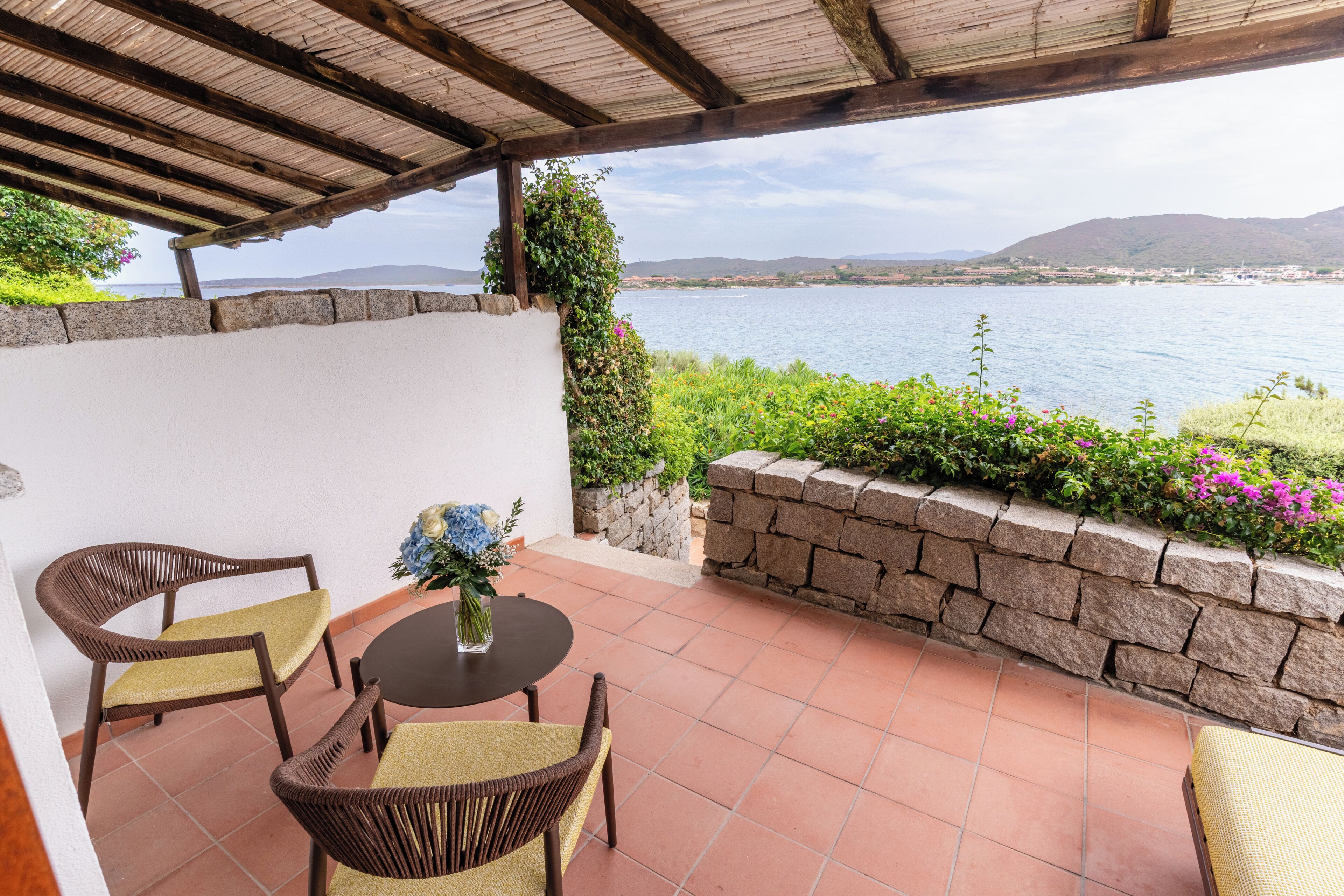 A covered patio with wicker furniture, a stone wall, and a view of the sea.