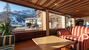 A wooden interior with a sofa and a table, large windows with a mountain view, and a plant.