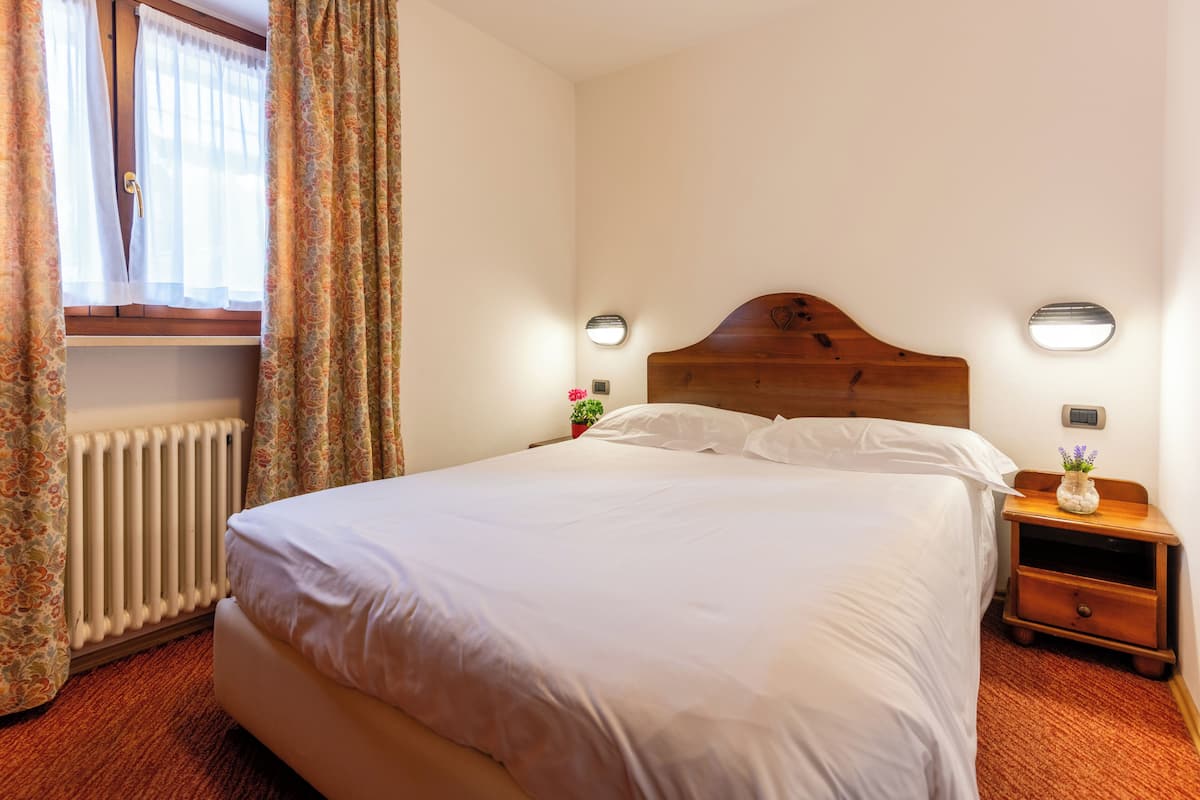 Una cama bien hecha con sábanas blancas, un cabecero de madera, una mesita de noche con una lámpara y una ventana con cortinas.