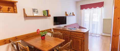 A dining area with a wooden table and chairs, a television on the wall, and a kitchen area with wooden cabinets and a countertop.