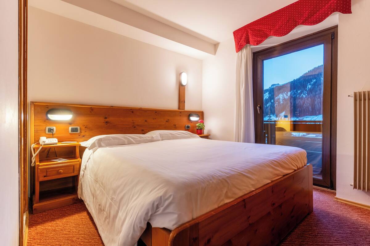 Un dormitorio con una cama de madera, una mesita de noche con un teléfono, una ventana que ofrece vistas a un paisaje nevado y una cortina roja.