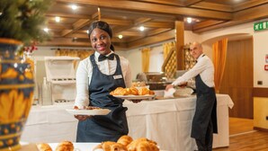 A buffet setup with a server holding a plate of pastries, a buffet table, and a counter with a person preparing food.