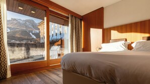 A bedroom with a large bed, wooden flooring, a sliding glass door leading to a balcony, and a mountain view.