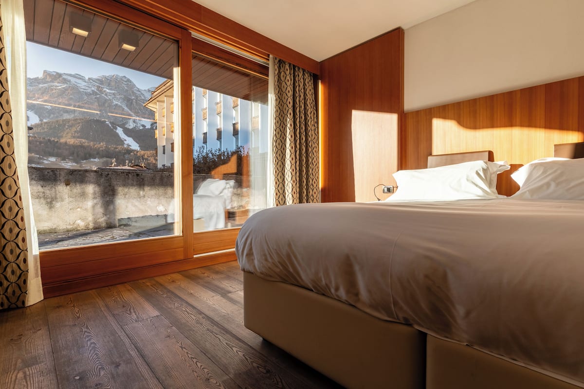 Un dormitorio con una cama grande, piso de madera, una puerta corrediza de vidrio hacia un balcón y vistas a la montaña.