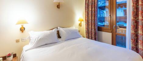 A hotel room with a large bed, two bedside lamps, a nightstand with a vase of flowers, and a window with curtains.