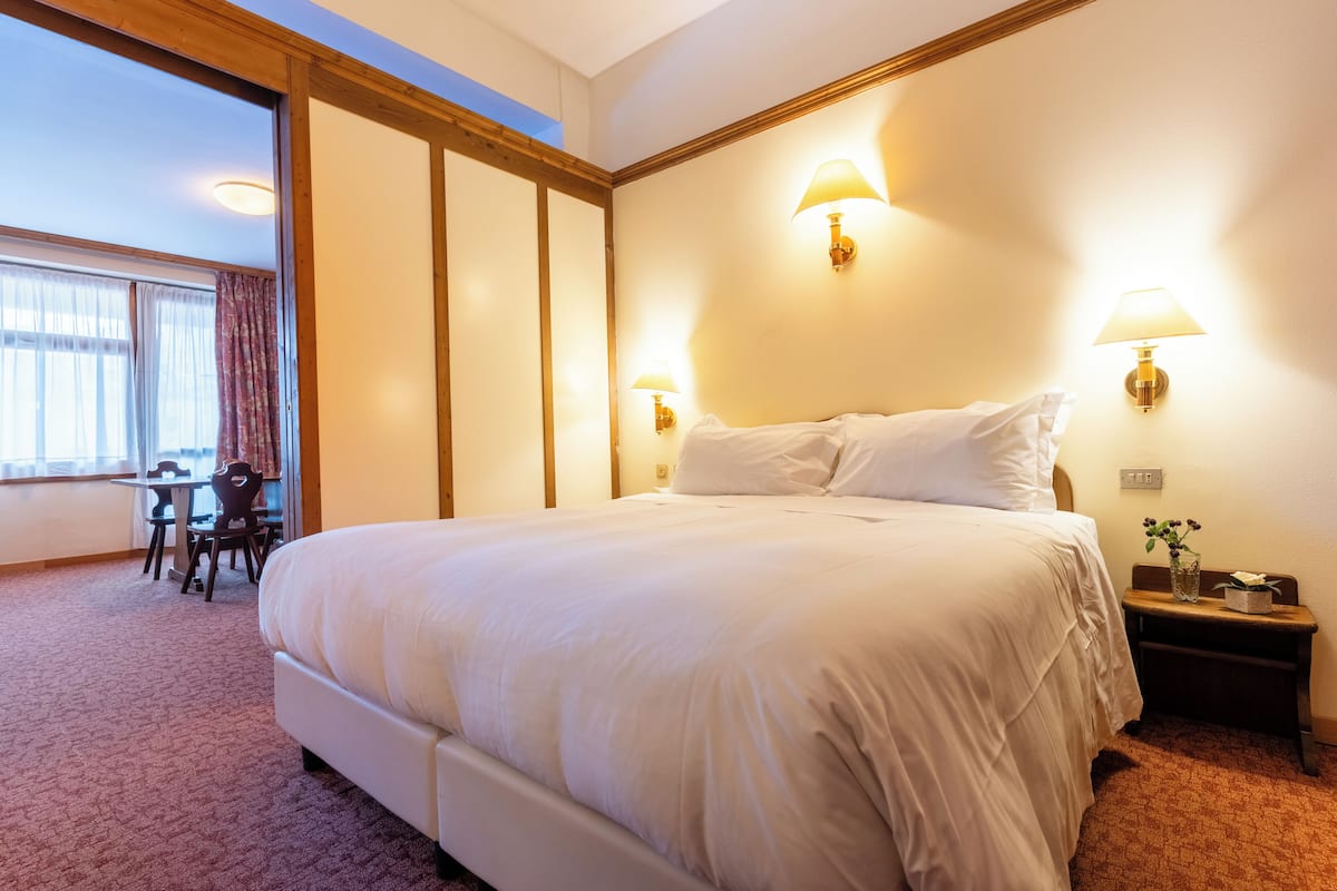 Una habitación de hotel con una cama grande, dos lámparas de noche, una mesita con una silla y un ventanal con cortinas.