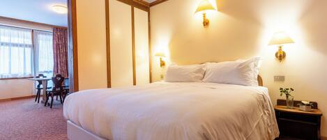 Una habitación de hotel con una cama grande, dos lámparas de noche, una mesita con una silla y un ventanal con cortinas.