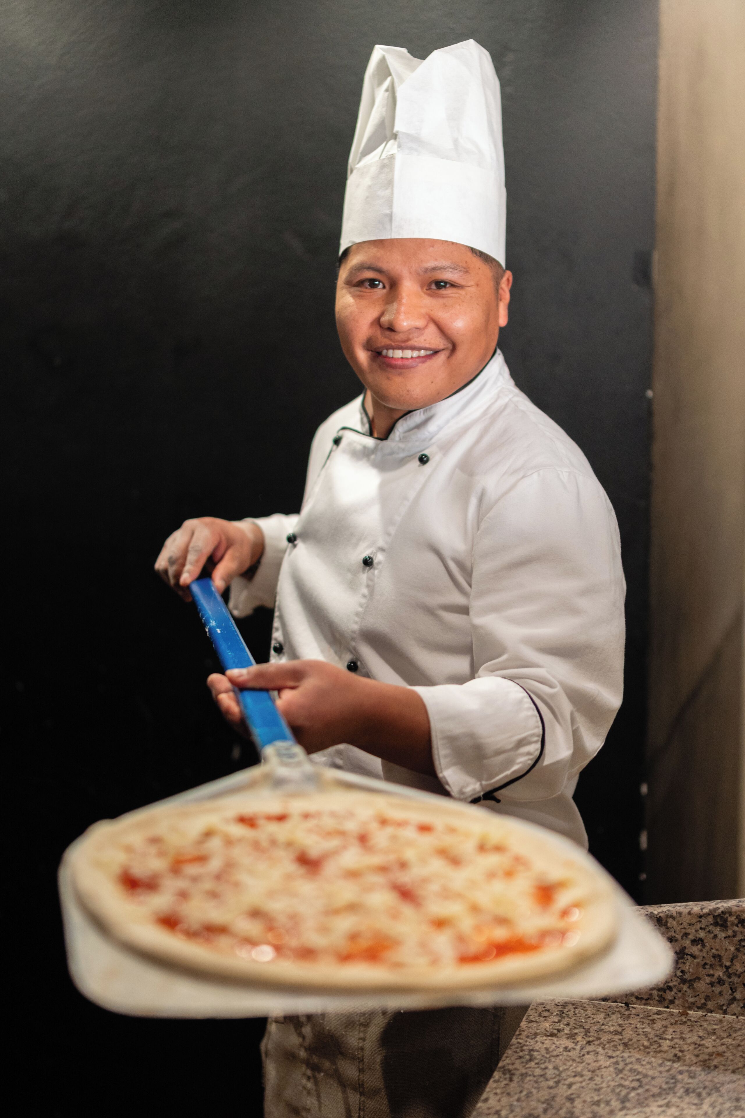 Un cocinero con uniforme blanco sostiene una pizza sobre una bandeja metálica.