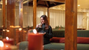A person sitting on a green sofa with a candle in the foreground, wooden beams, and a window with curtains in the background.