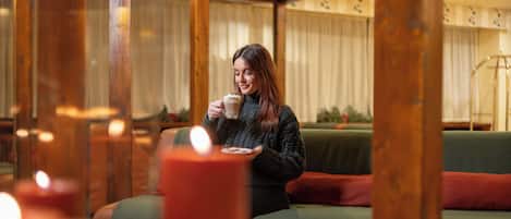 A person sitting on a green sofa with a candle in the foreground, wooden beams, and a window with curtains in the background.