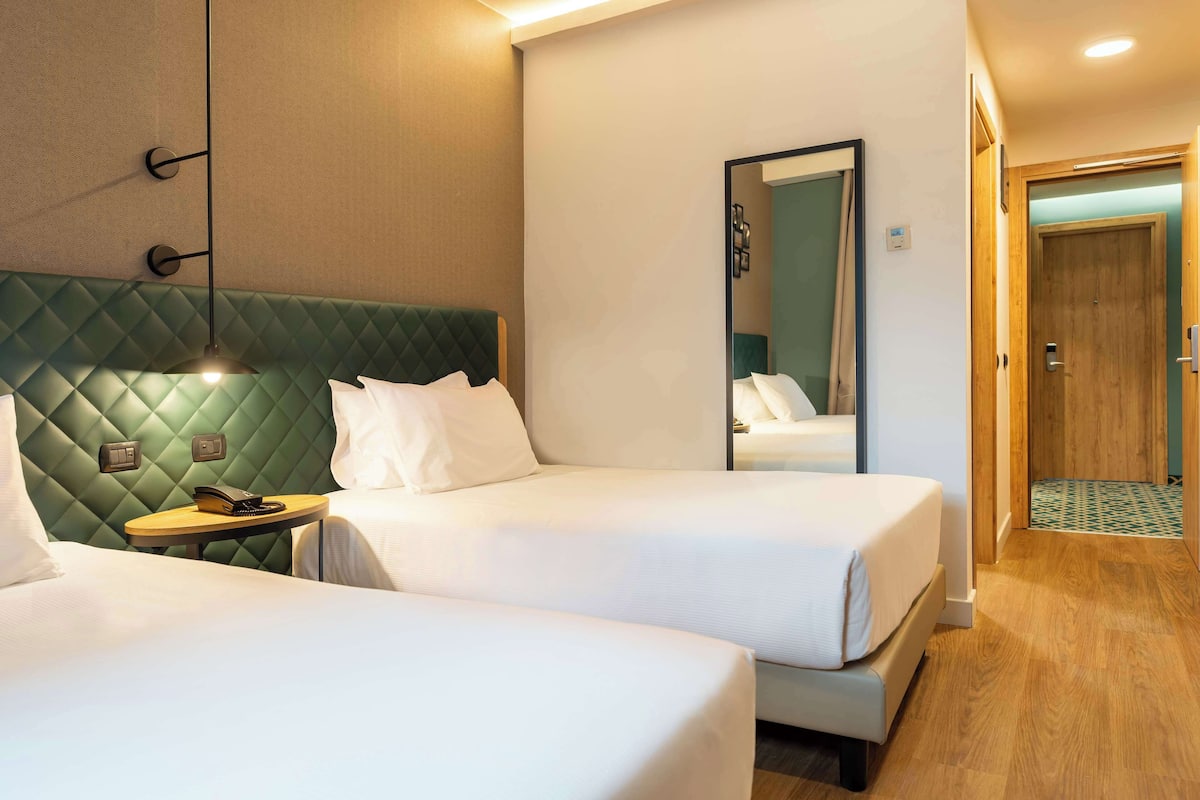 A hotel room with two beds, a green quilted headboard, a bedside table with a lamp, and a mirror on the wall.