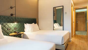 Habitación de hotel con dos camas, cabecera verde acolchada, mesita de noche con lámpara y espejo en la pared.