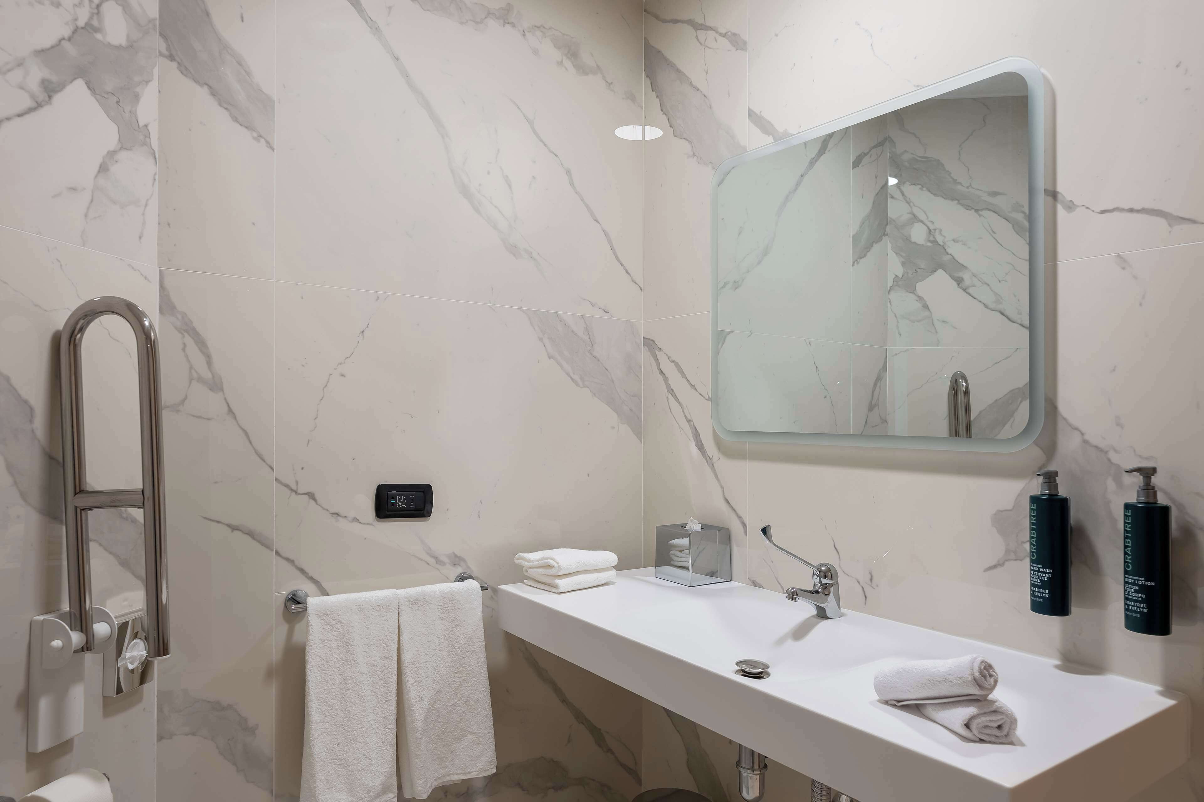 A bathroom with a marble wall, a white sink, a towel rack, a glass-topped vanity, and two bottles of hand soap.