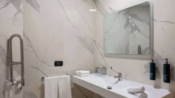 A bathroom with a marble wall, a white sink, a towel rack, a glass-topped vanity, and two bottles of hand soap.