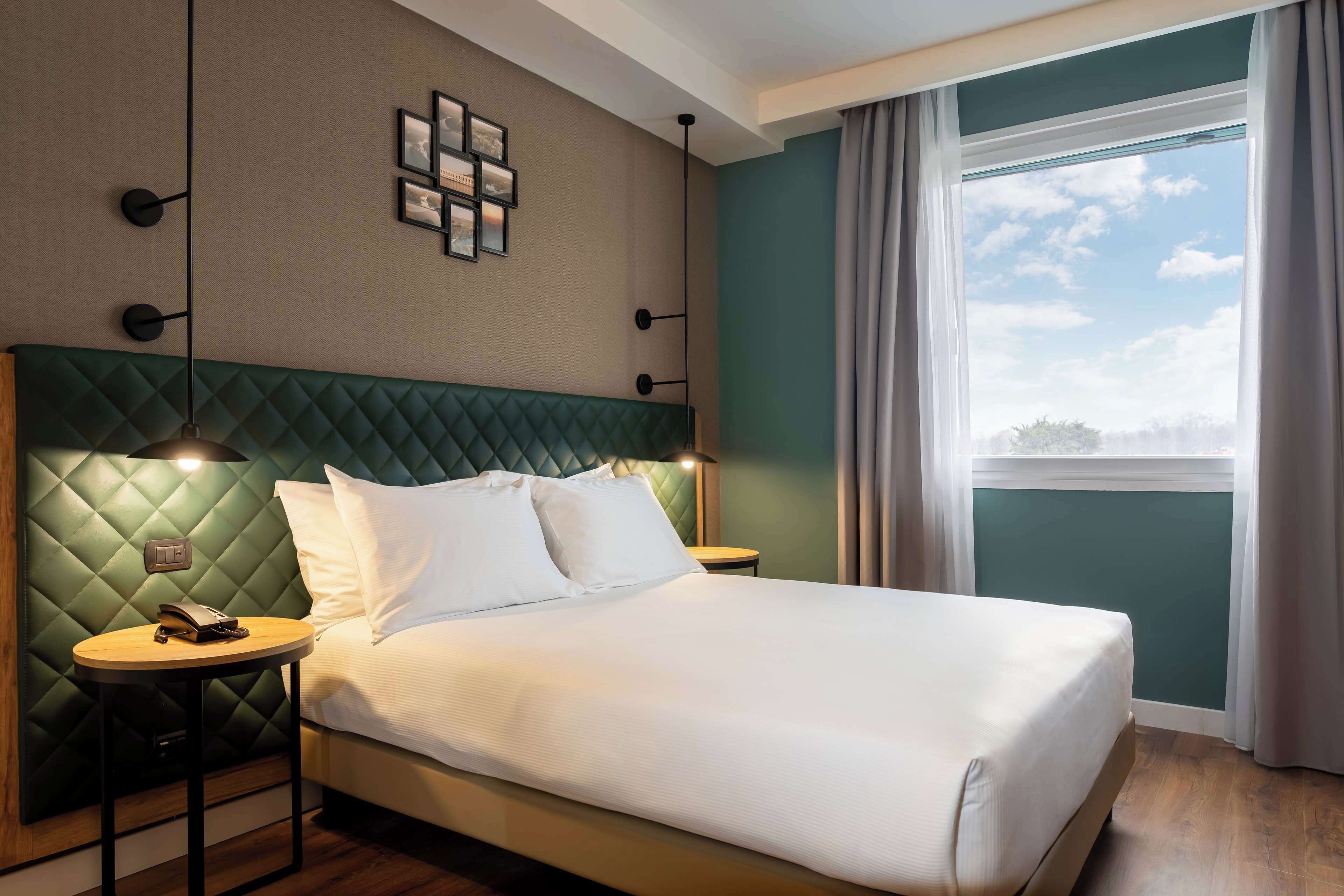 Una cama bien hecha con cabecera verde, dos mesitas de noche con lámparas y vista al cielo a través de una ventana.