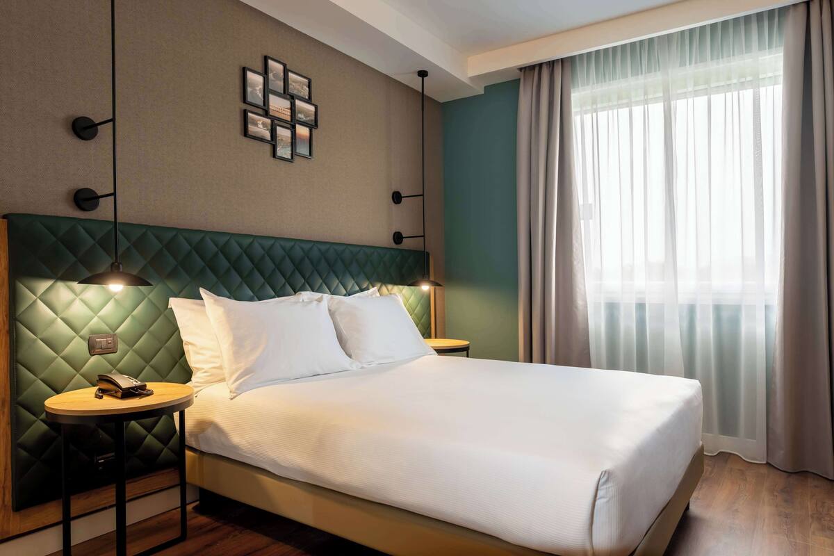 A neatly made bed with a green headboard, a bedside table with a lamp, and a window with sheer curtains.