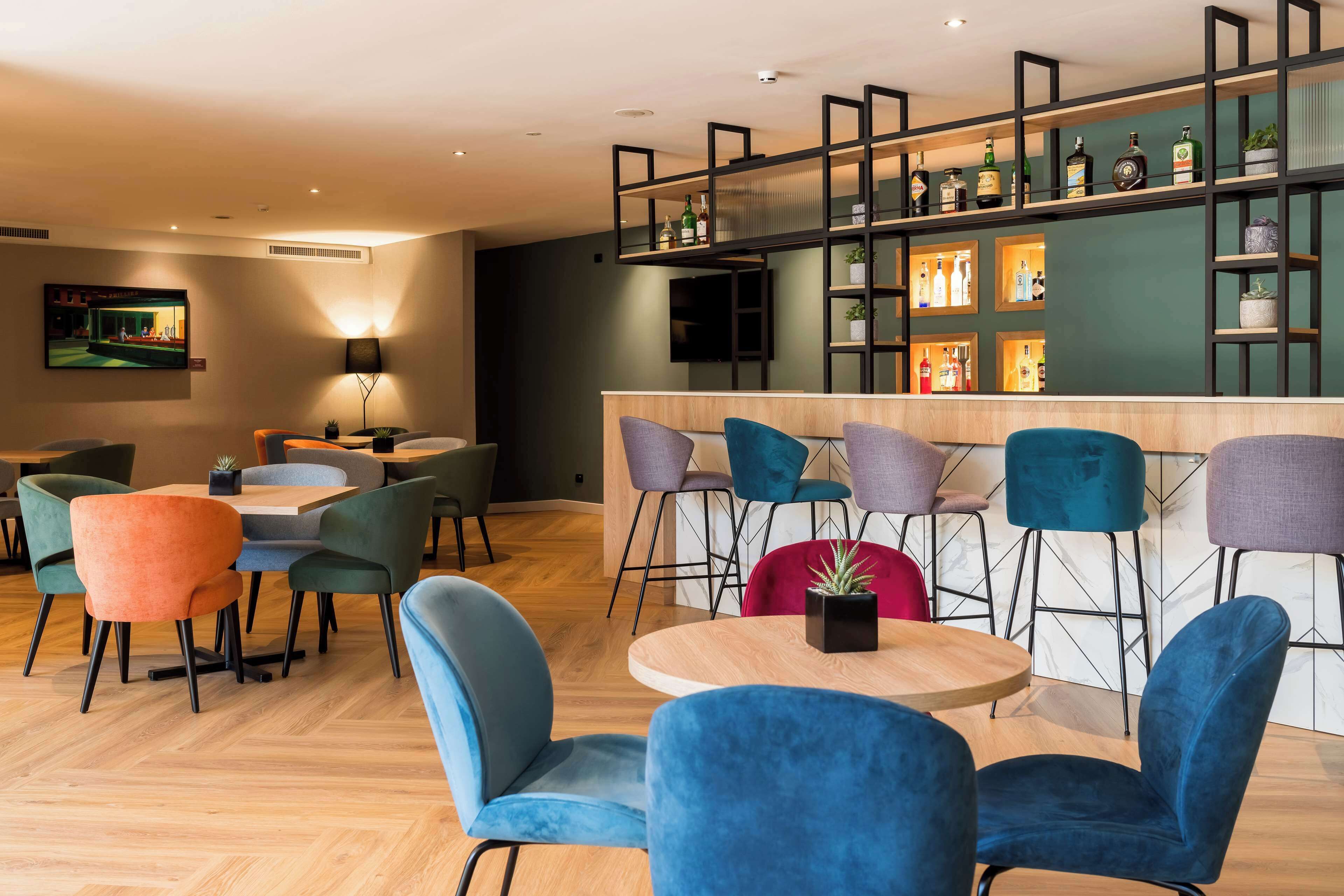 A modern bar area with a variety of seating options including round tables and bar stools, a television mounted on the wall, and shelves displaying bottles and plants.