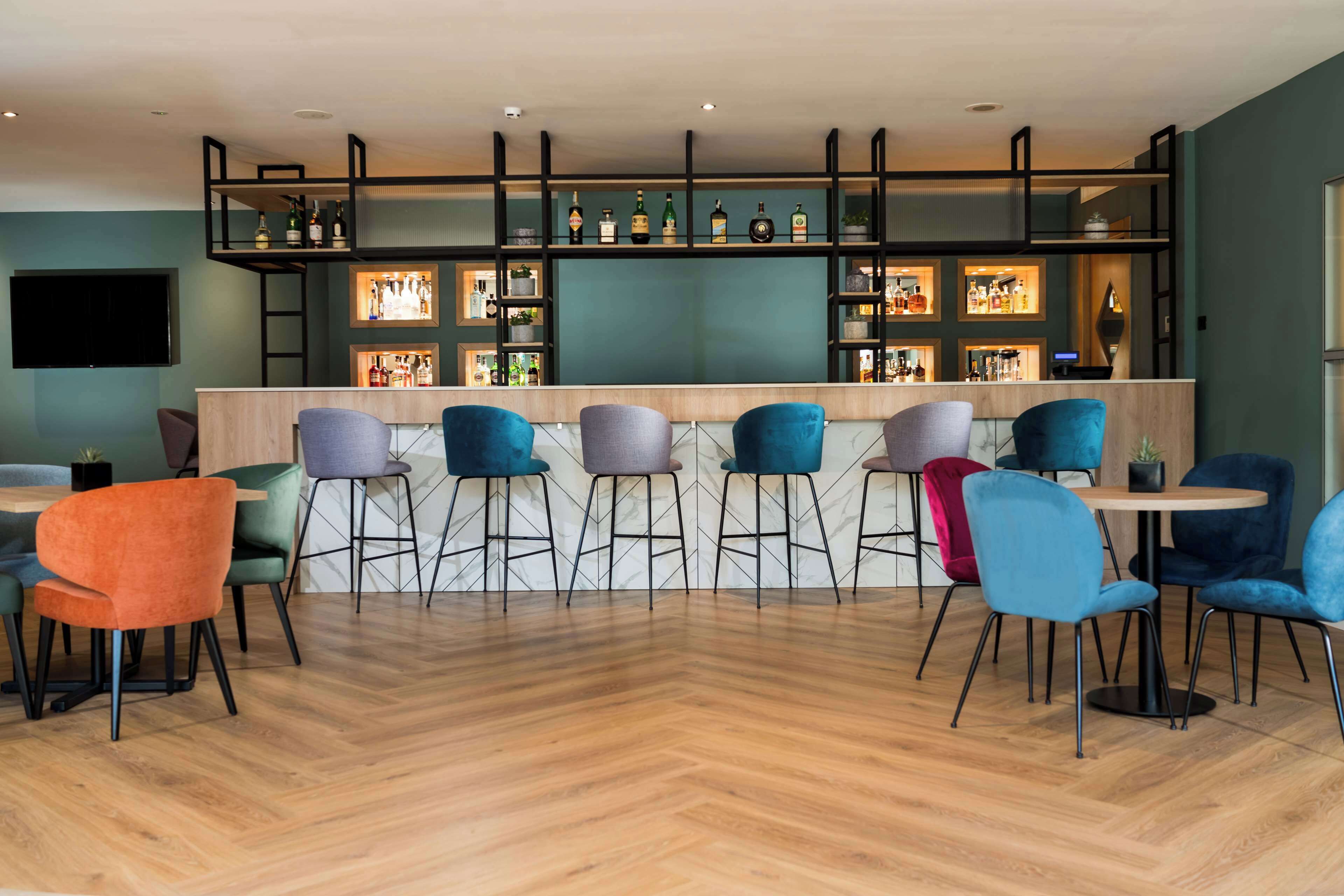 A bar area with a wooden floor, multiple stools, and a variety of bottles on shelves.