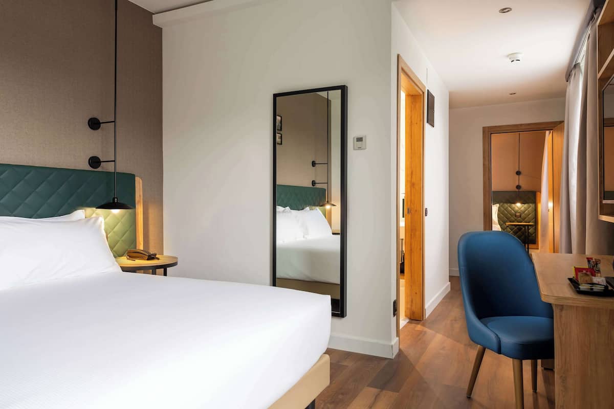 A hotel room with a bed, a desk, a chair, a mirror, and a door leading to another room.