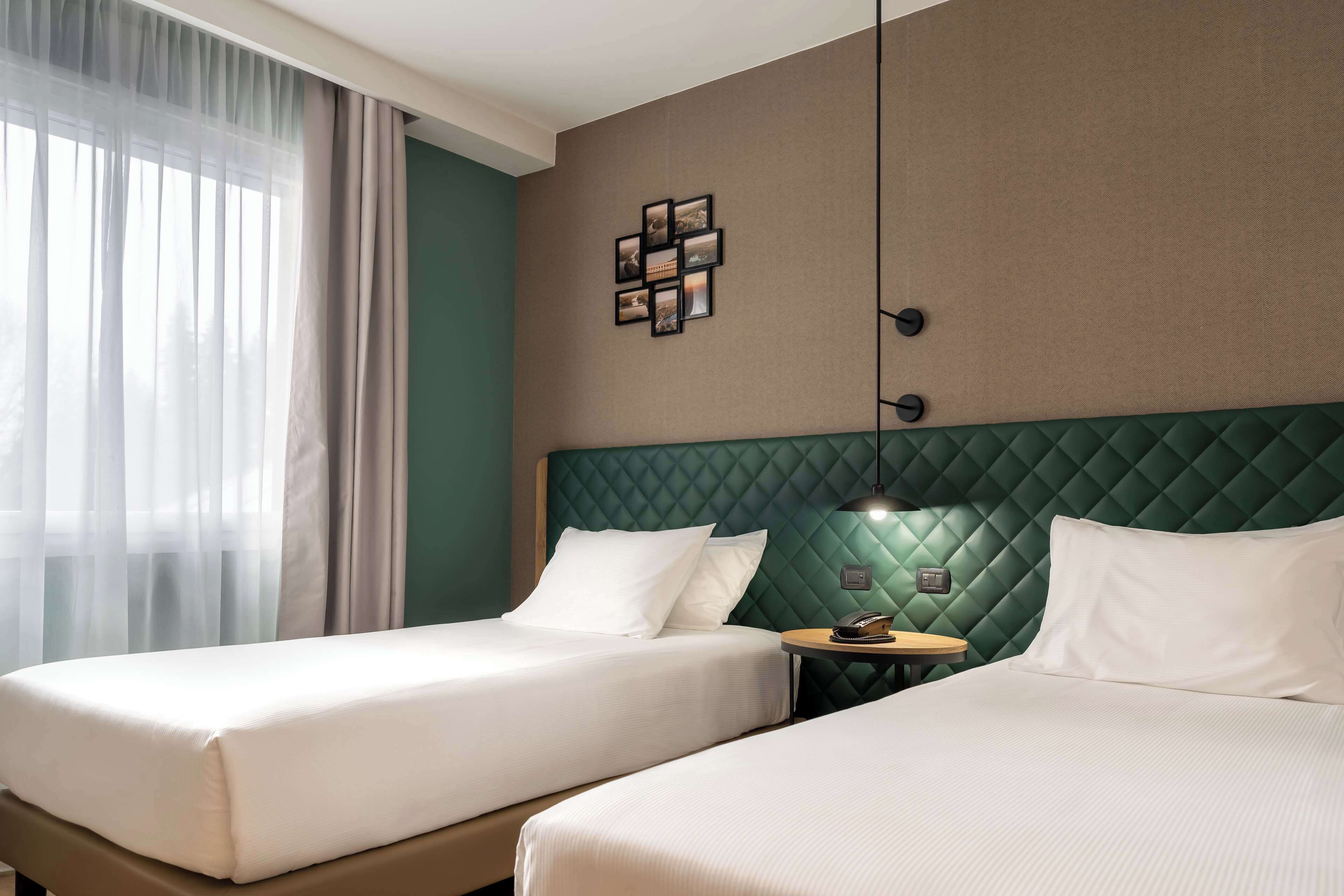 Una habitación de hotel con dos camas, cabecera verde, lámpara de pared y una mesita con un teléfono.