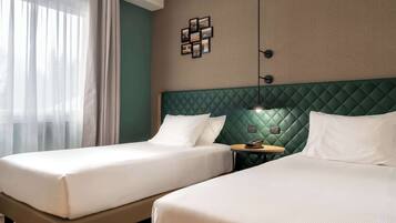 Una habitación de hotel con dos camas, cabecera verde, lámpara de pared y una mesita con un teléfono.