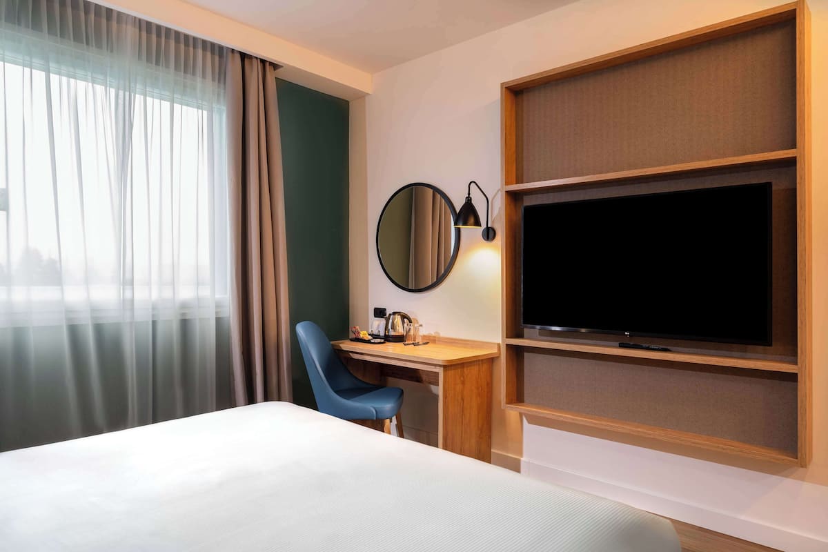 A hotel room with a bed, a desk with a chair, a television, and a large mirror.