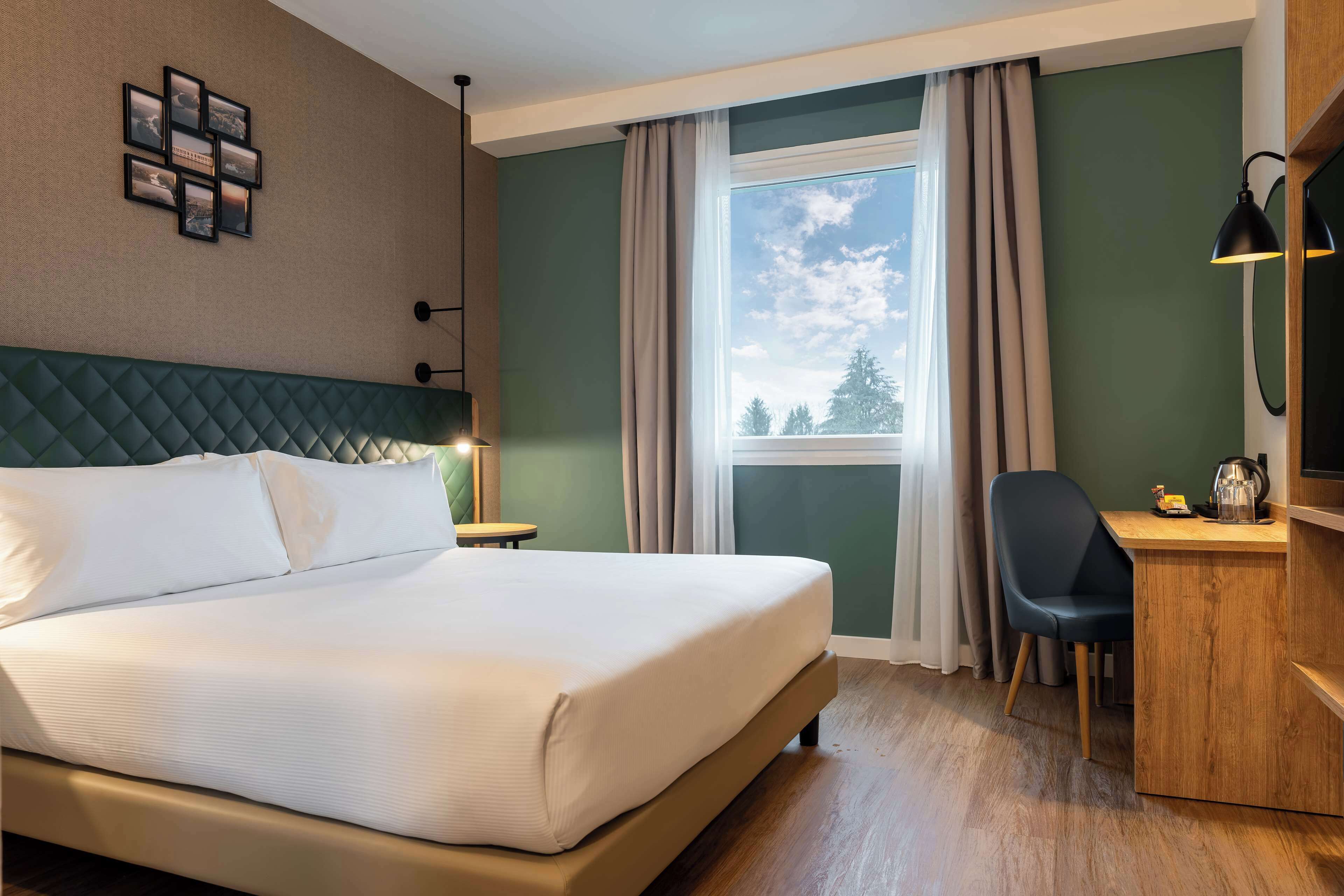 A hotel room with a bed, a desk, a chair, a window with curtains, a wall-mounted picture, and a lamp.