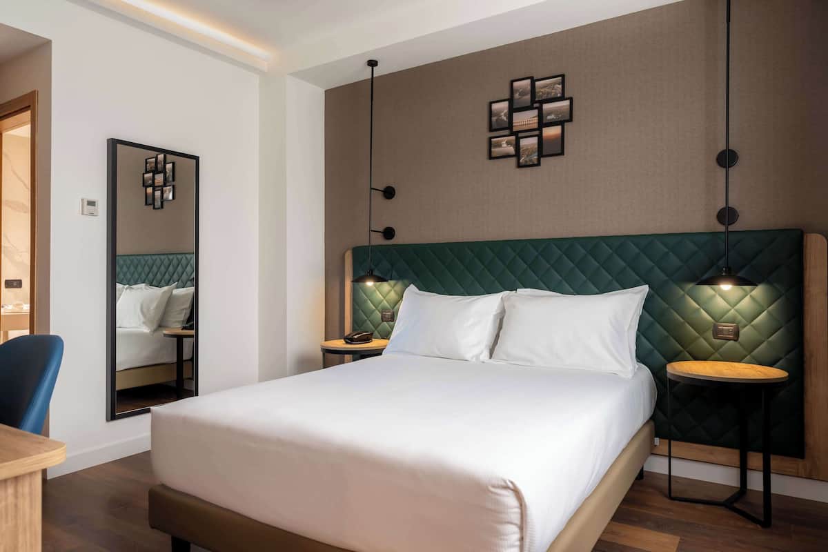 A hotel room with a large bed, a mirror, a desk, and a green headboard.