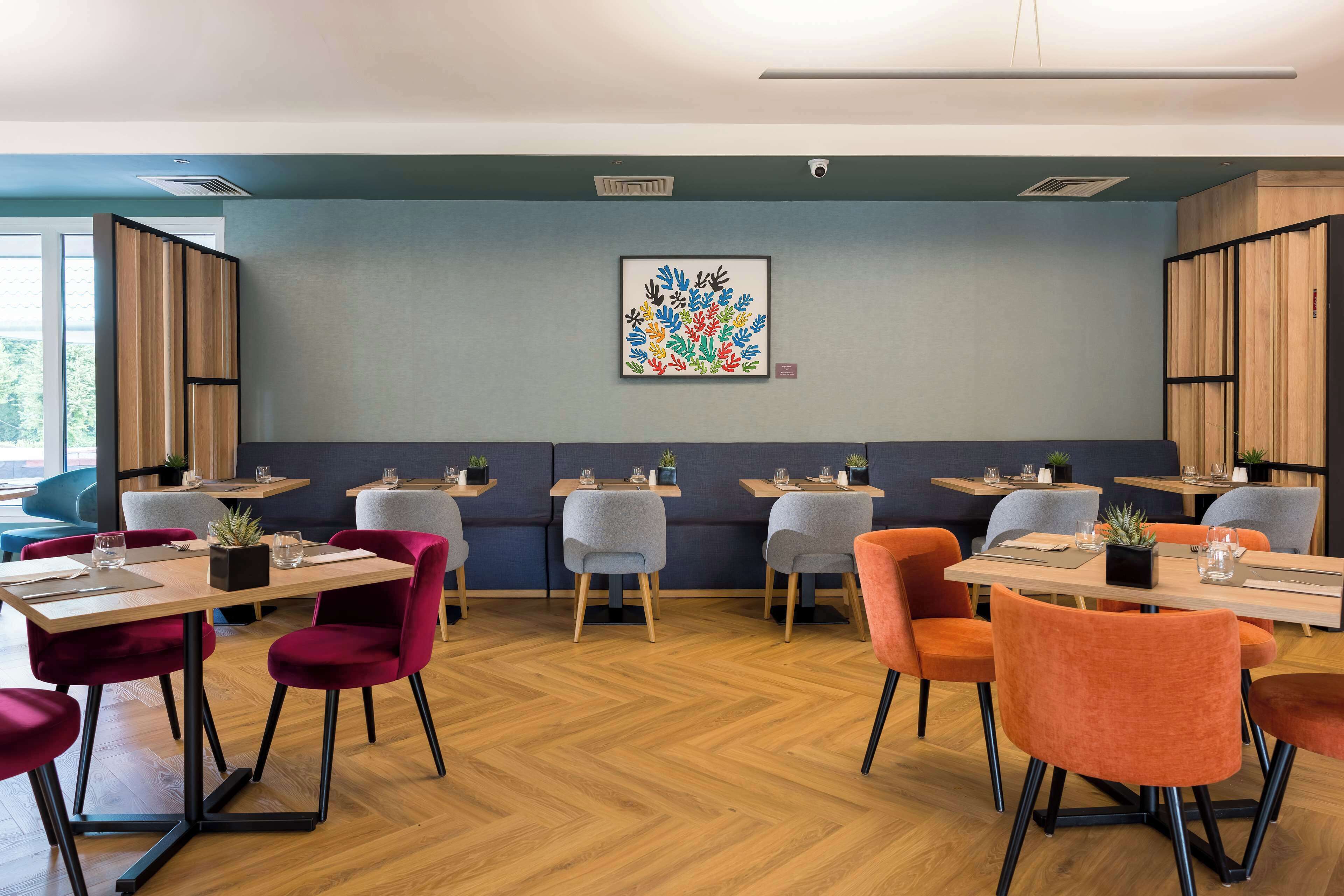 Un comedor moderno con mesas de madera, sillas tapizadas en rojo y gris, y una pared azul decorada con una obra de arte colorida.
