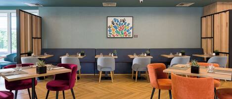 Un comedor moderno con mesas de madera, sillas tapizadas en rojo y gris, y una pared azul decorada con una obra de arte colorida.
