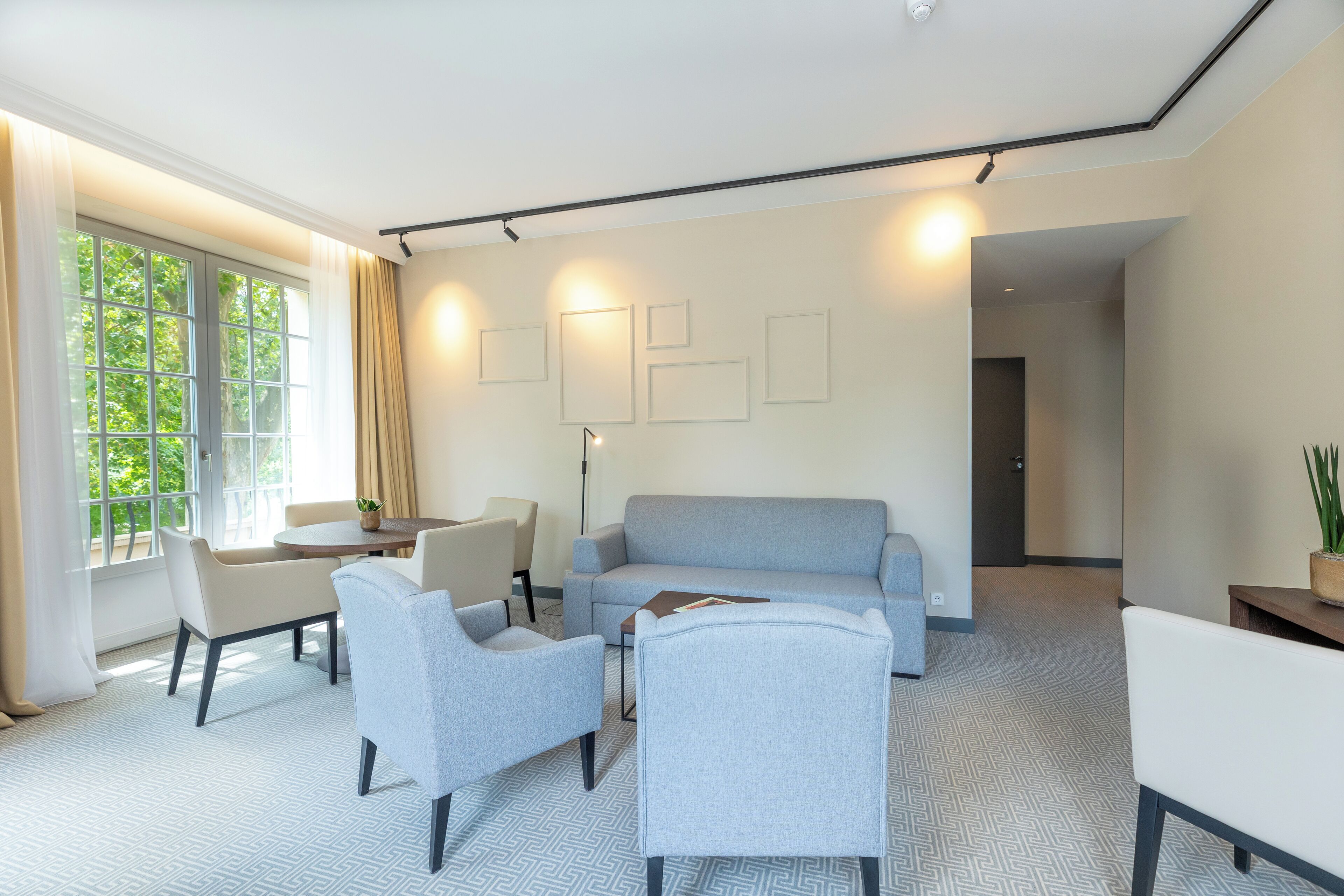Ein modernes Wohnzimmer mit einem grauen Sofa, zwei Sesseln, einem Couchtisch und einem Esstisch. An der Wand hängen gerahmte Bilder, ein großes Fenster mit Vorhängen und eine Tür, die in einen anderen Raum führt.