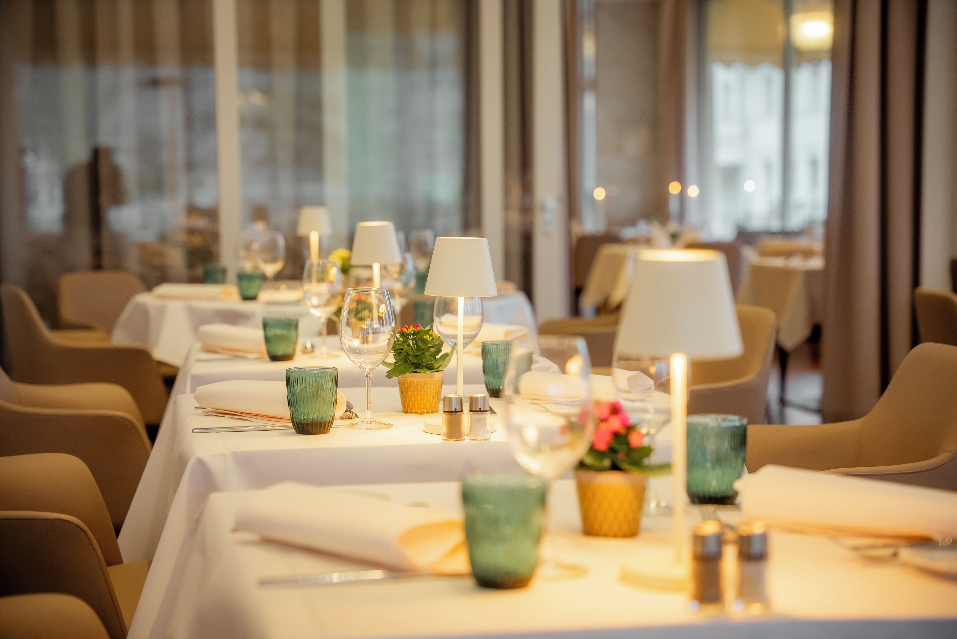 A restaurant with tables set for dining, featuring candles, glassware, and small potted plants.