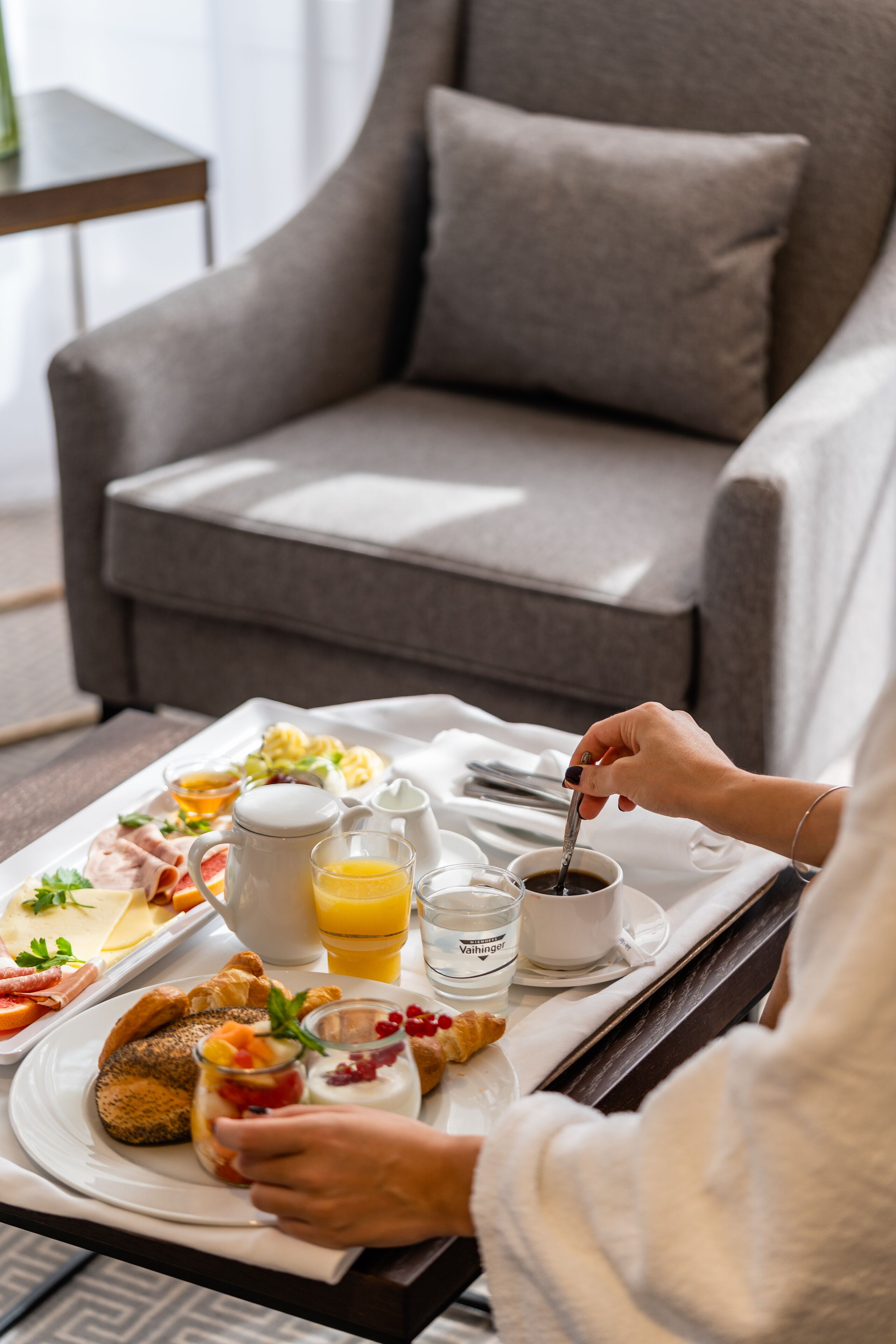 Bandeja de desayuno con croissants, queso y fruta sobre una mesa en una habitación con un sofá gris y una pequeña mesa al fondo.