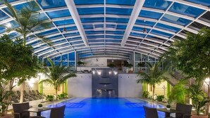 An indoor swimming pool with a glass ceiling, surrounded by potted plants and outdoor seating. - Parkhotel Kurhaus (Bad Kreuznach)
