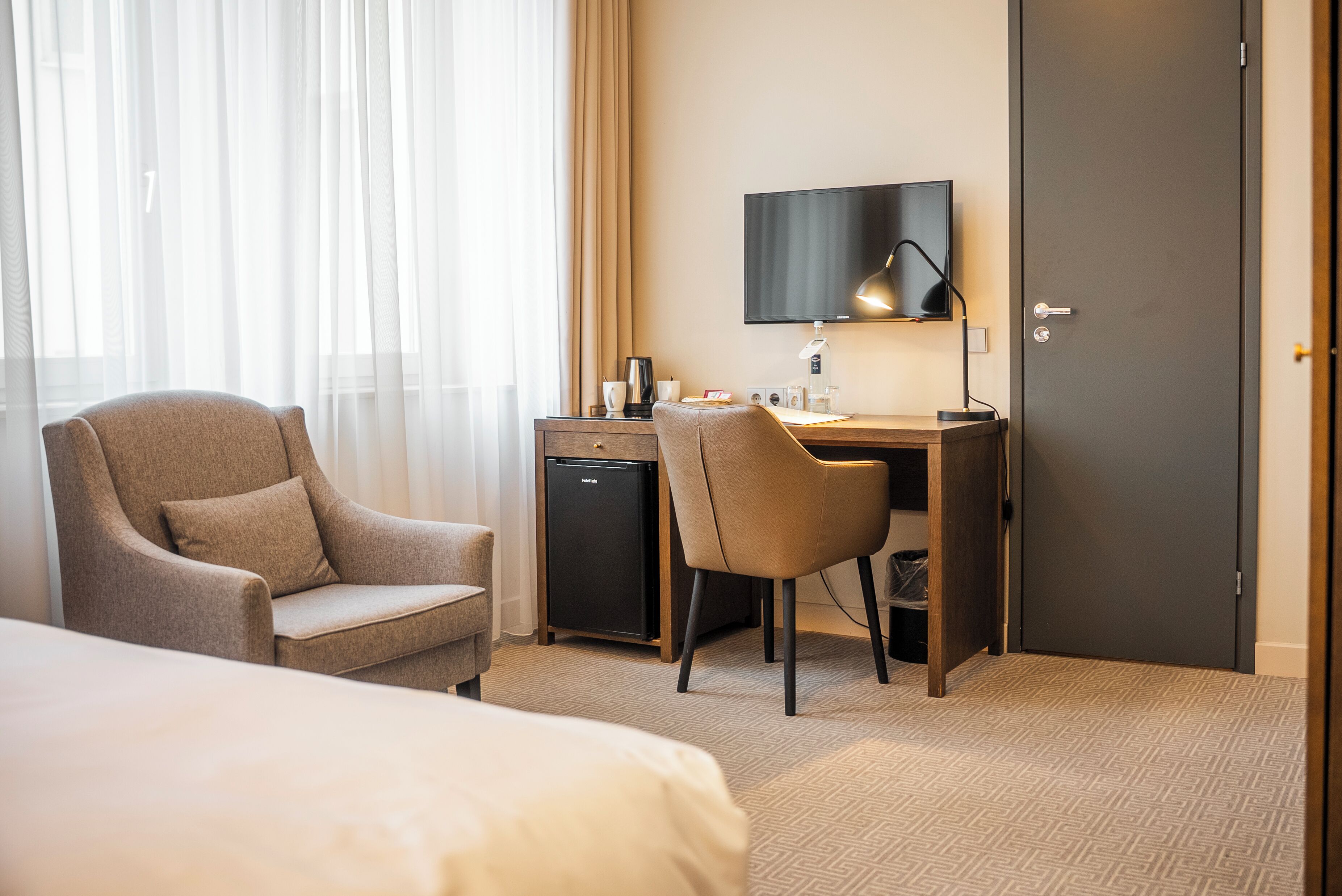 Een hotelkamer met een bed, een bureau met een computer, een stoel, een televisie en een minibar.