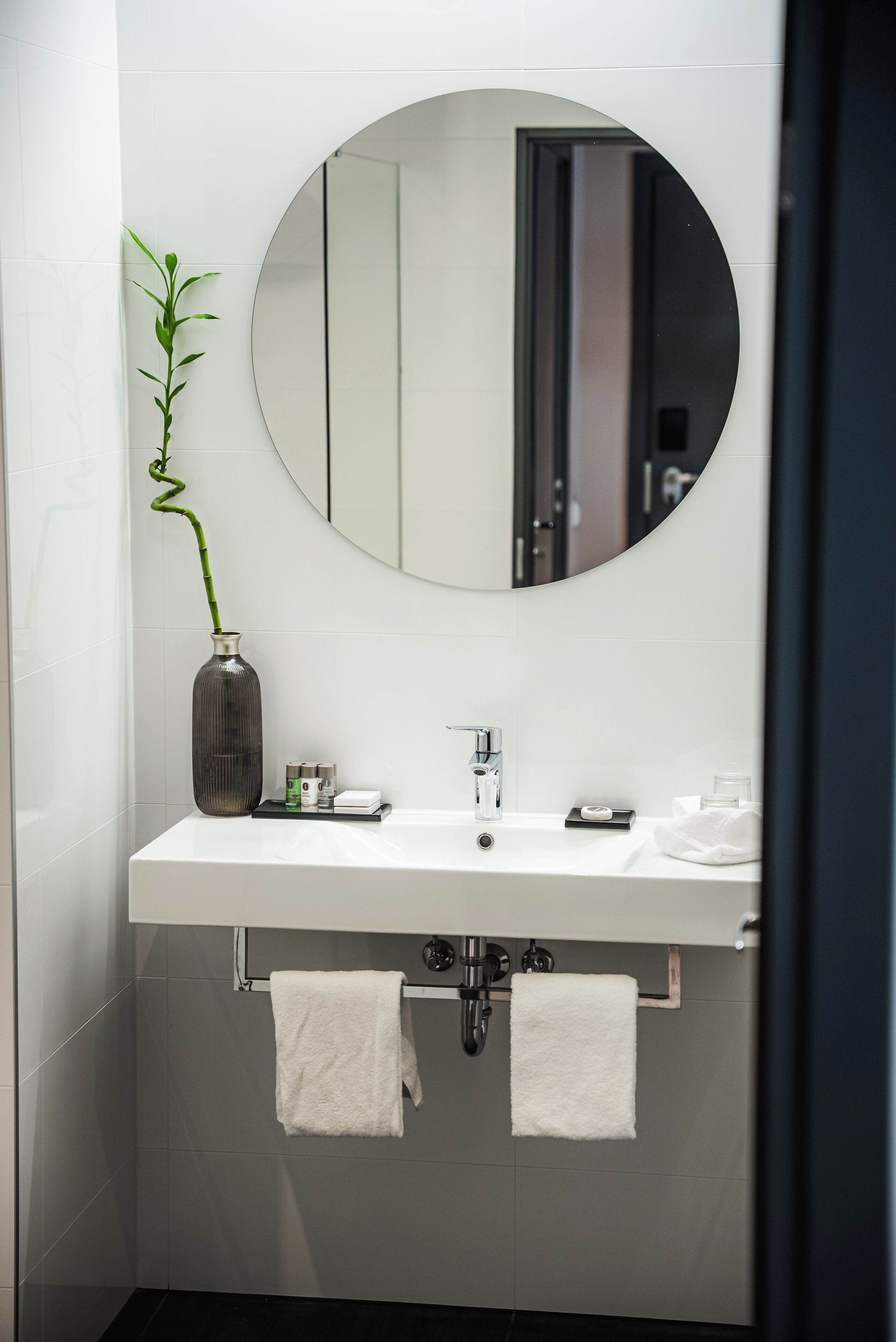 Un baño moderno con un lavamanos blanco, un espejo redondo y una planta de bambú en un florero.