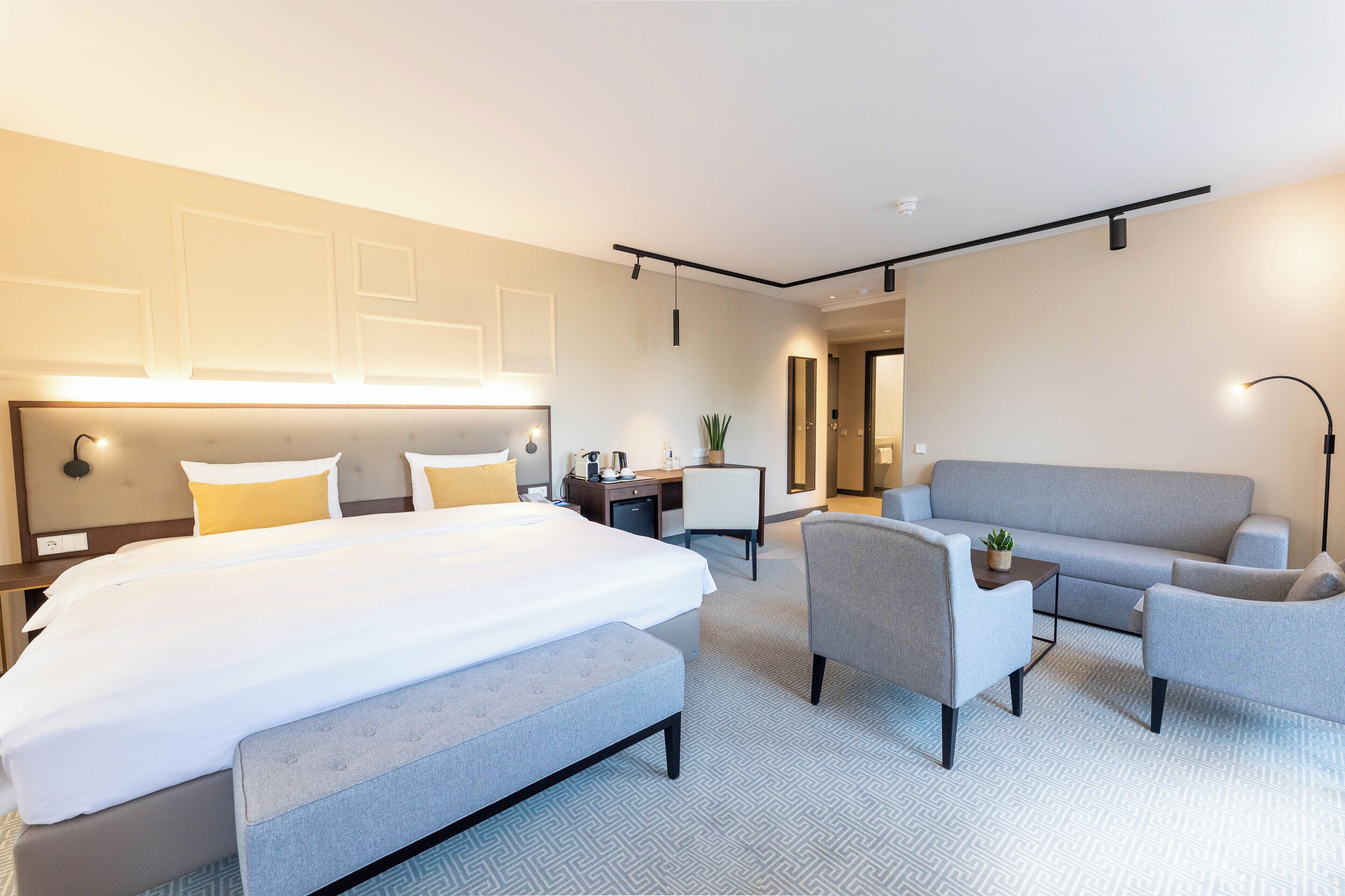 A modern hotel room with a large bed, a sofa, two armchairs, a desk, and a small plant.